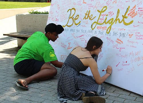 Students sitting on the ground writing a message on a board that says Be the Light