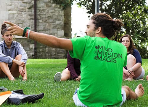 An older student talking to new freshman sitting in a circle on a grassy field