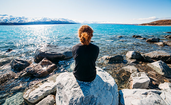 Student sitting on a rock, admiring a glassy blue body of water with mountains in the distance