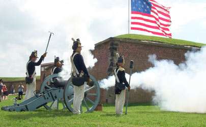 A cannon surrounded by reenactors dressed in military uniforms fires at Fort McHenry in Baltimore